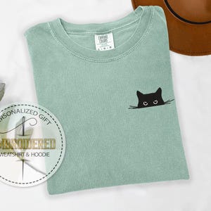 May include: A light green t-shirt with a black cat face embroidered on the front. The cat is peeking out from behind a line, with only its eyes and ears visible. The t-shirt has a crew neck and short sleeves.