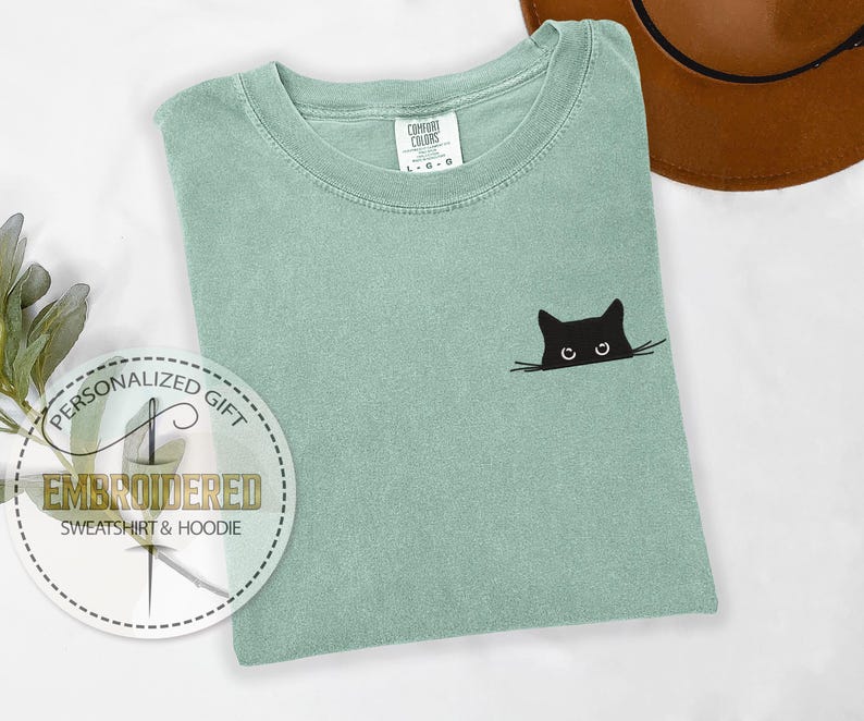 May include: A light green t-shirt with a black cat face embroidered on the front. The cat is peeking out from behind a line, with only its eyes and ears visible. The t-shirt has a crew neck and short sleeves.
