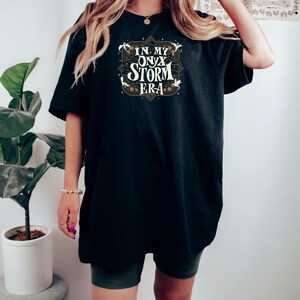 May include: Black t-shirt with the text "IN MY ONYX STORM ERA" in a decorative design. The text is in white and gold. The shirt is a loose fit.