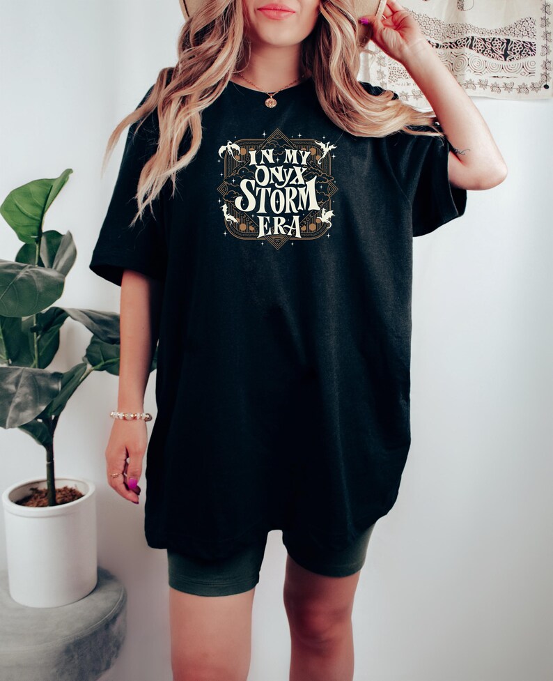 May include: Black t-shirt with the text "IN MY ONYX STORM ERA" in a decorative design. The text is in white and gold. The shirt is a loose fit.