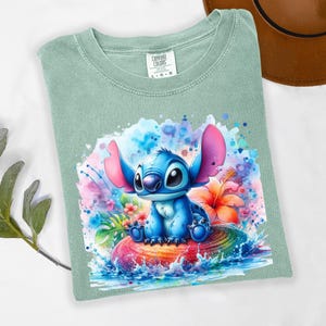 Może przedstawiać: Szałwiowa koszulka Comfort Colors z akwarelową ilustracją Stitcha z Lilo & Stitch. Projekt przedstawia Stitcha siedzącego na kolorowej, tęczowej powierzchni z tropikalnymi kwiatami i rozpryskami wody.