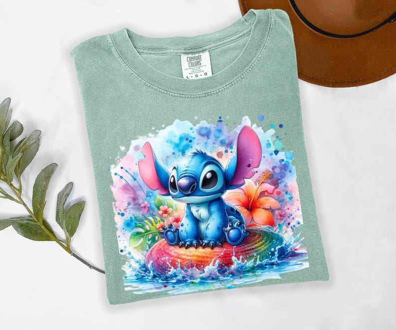 Może przedstawiać: Szałwiowa koszulka Comfort Colors z akwarelową ilustracją Stitcha z Lilo & Stitch. Projekt przedstawia Stitcha siedzącego na kolorowej, tęczowej powierzchni z tropikalnymi kwiatami i rozpryskami wody.