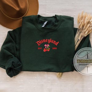 May include: A dark green sweatshirt with a red embroidered design that says "Disneyland Est. 1955" with a red cherry and pink bow.