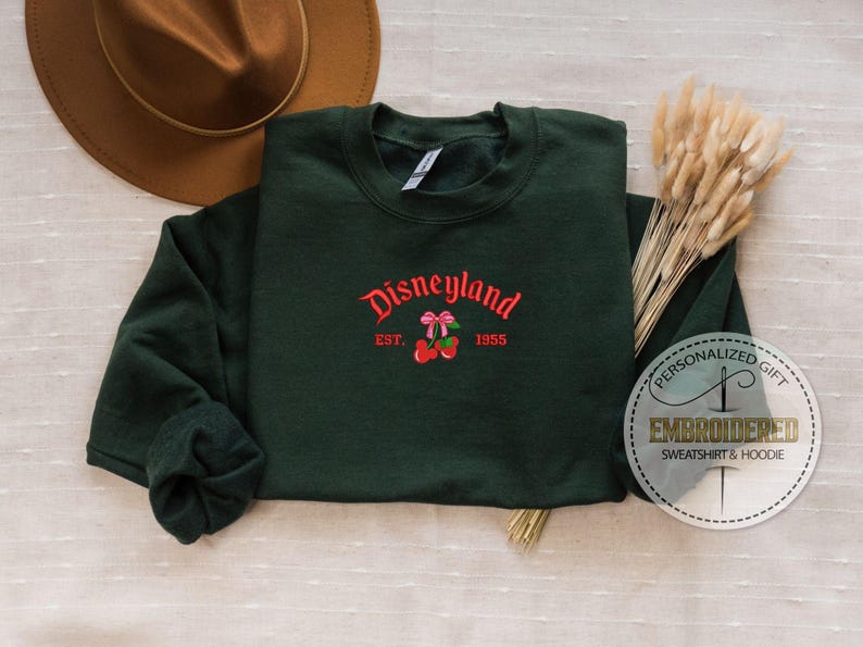 May include: A dark green sweatshirt with a red embroidered design that says "Disneyland Est. 1955" with a red cherry and pink bow.