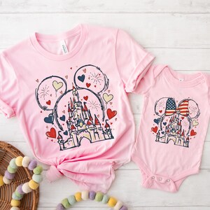 May include: Pink t-shirts and a onesie featuring a castle design within Mickey Mouse ears, with hearts and fireworks. The onesie has an American flag bow. The t-shirts are a matching set.