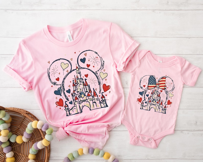 May include: Pink t-shirts and a onesie featuring a castle design within Mickey Mouse ears, with hearts and fireworks. The onesie has an American flag bow. The t-shirts are a matching set.