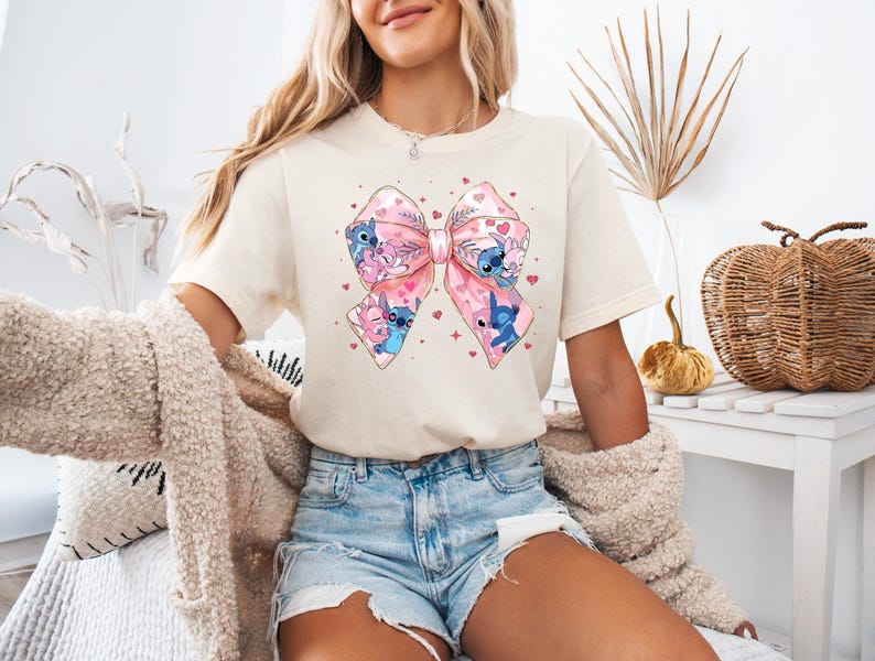 May include: A white t-shirt with a pink and blue bow featuring cartoon characters. The bow is decorated with hearts and flowers.