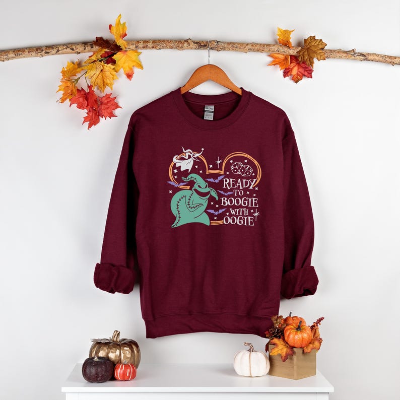 Pu&ograve; includere: Felpa bordeaux con una grafica di Oogie Boogie, pipistrelli e la scritta "Ready to Boogie with Oogie!". Il design &egrave; nei toni del verde, viola e arancione. La felpa &egrave; appesa a un gancio di legno.