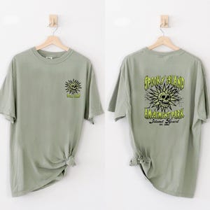 May include: Two sage green t-shirts are displayed. One shows a sun-like skull graphic with the text "Spooky Island" on the front. The other shirt has the text "Spooky Island Amusement Park Island Resort Est. 2002" with a skull graphic.