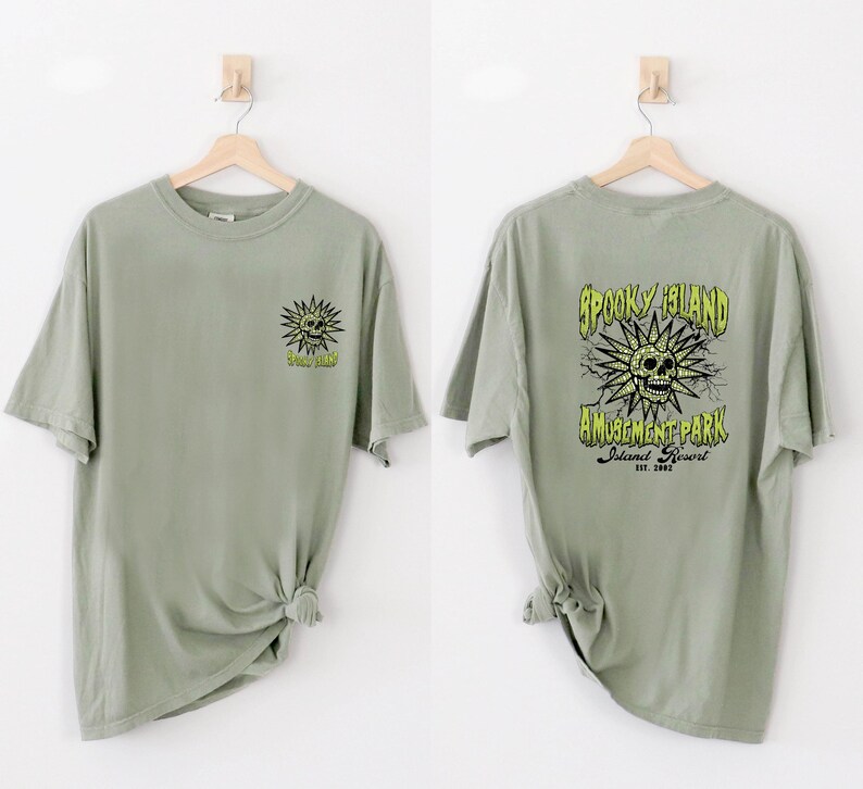 May include: Two sage green t-shirts are displayed. One shows a sun-like skull graphic with the text "Spooky Island" on the front. The other shirt has the text "Spooky Island Amusement Park Island Resort Est. 2002" with a skull graphic.