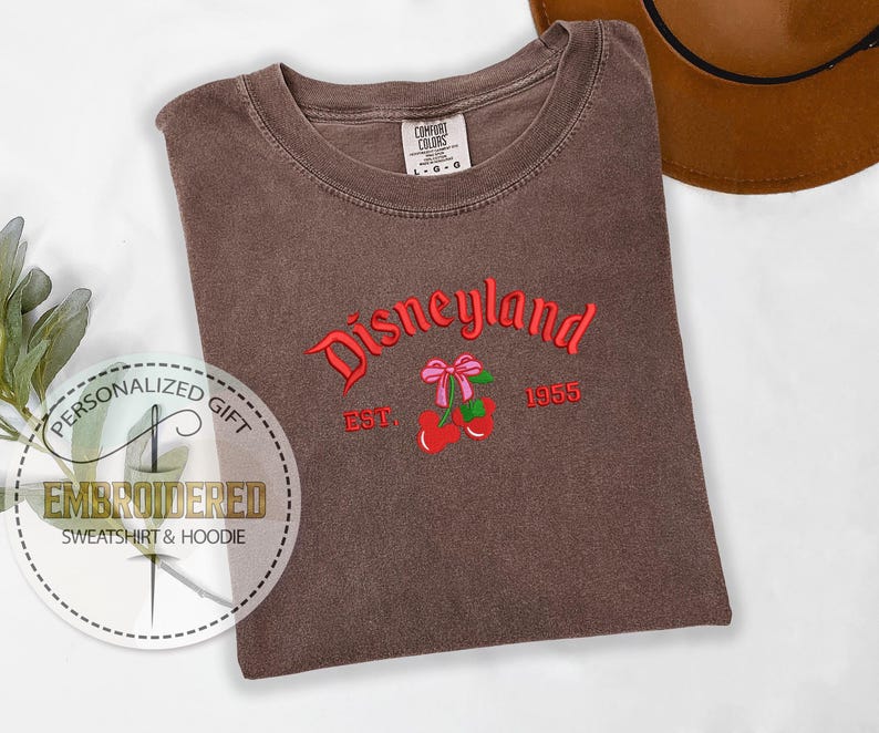 Op de afbeelding: Bruin T-shirt met de tekst "Disneyland Est. 1955" geborduurd in rood. De tekst is gebogen en heeft aan elke kant een rode kers met een roze strik.
