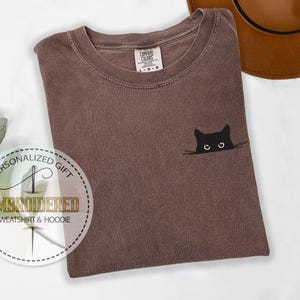 May include: A brown t-shirt with a black cat peeking out from behind a white line. The cat has white eyes and a black nose.