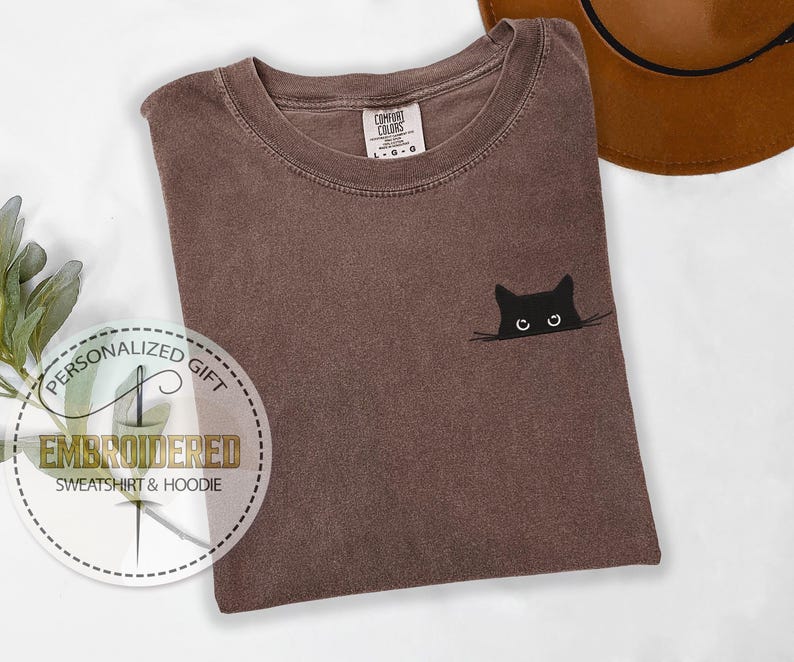 May include: A brown t-shirt with a black cat peeking out from behind a white line. The cat has white eyes and a black nose.