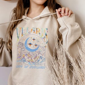 May include: Beige hoodie with a colorful graphic design. The design features the word "VELARIS" in a rainbow font above a crescent moon, stars, and mountains. The text "CITY OF STARLIGHT" is below the graphic.