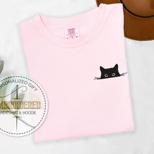 May include: A pink t-shirt with a black embroidered cat peeking out from behind the fabric. The cat has large eyes and a small nose.
