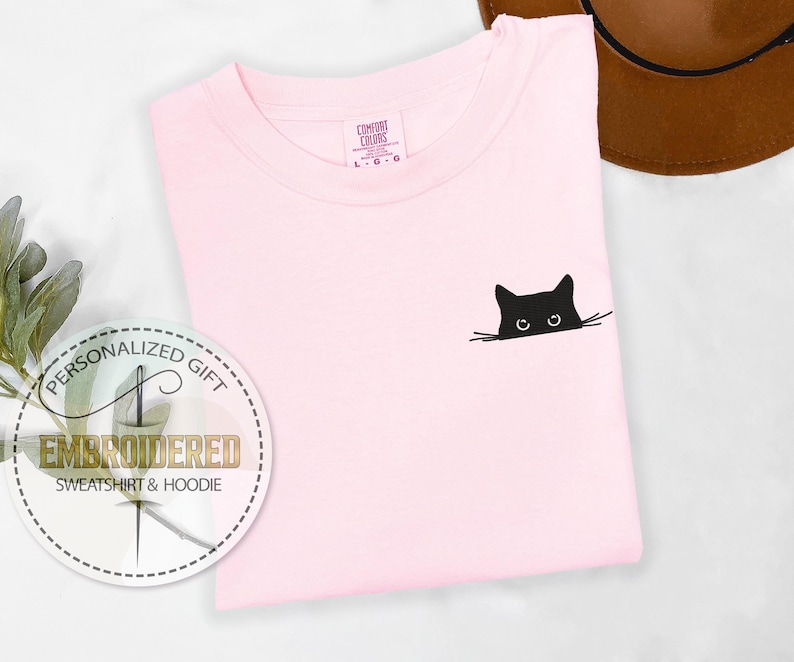 May include: A pink t-shirt with a black embroidered cat peeking out from behind the fabric. The cat has large eyes and a small nose.