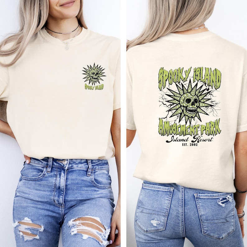 May include: Cream-colored t-shirt with a front and back graphic. The front features a green and black sunburst design with a skull and the words "Spooky Island". The back has a larger version of the design with "Amusement Park Island Resort EST. 2002".