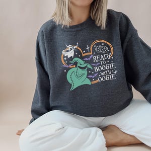 Pu&ograve; includere: Felpa grigio scuro con un design a forma di cuore e la scritta "READY TO BOOGIE WITH OOGIE". Il design include un personaggio verde di Oogie Boogie, dadi, pipistrelli e un fantasma. La felpa &egrave; uno stile casual e confortevole.