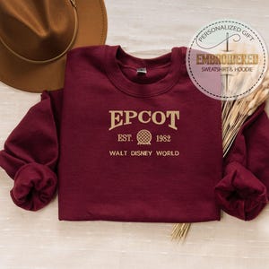 Puede incluir: Una sudadera granate con la palabra "EPCOT" en letras doradas, junto con "EST. 1982" y un dise&ntilde;o de globo. Las palabras "WALT DISNEY WORLD" est&aacute;n debajo. Un sombrero marr&oacute;n est&aacute; en el fondo.