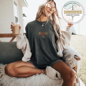 May include: A dark gray t-shirt with a red, yellow, and black embroidered design of Mickey Mouse. The shirt is worn by a person sitting on a white fluffy chair.