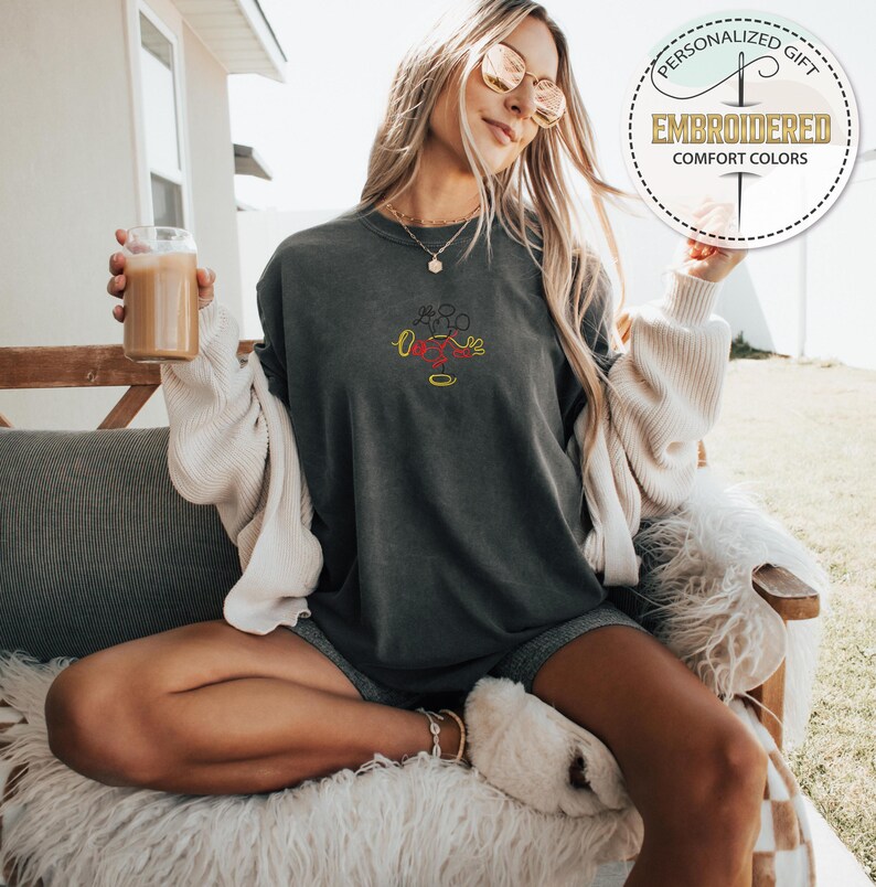 May include: A dark gray t-shirt with a red, yellow, and black embroidered design of Mickey Mouse. The shirt is worn by a person sitting on a white fluffy chair.