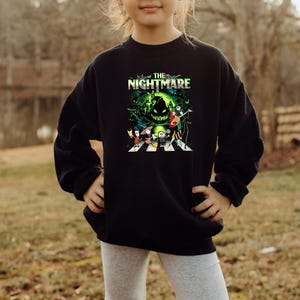 May include: Black sweatshirt featuring a colorful graphic from "The Nightmare Before Christmas." The design includes characters like Jack Skellington and Sally, with the title text "The Nightmare" in green and white.