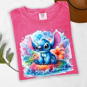 Może przedstawiać: R&oacute;żowa koszulka Comfort Colors z akwarelową ilustracją postaci Stitch. Niebieski Stitch siedzi na kolorowej, tęczowej powierzchni z wodą i kwiatami wok&oacute;ł niego. Koszulka w rozmiarze L-G.