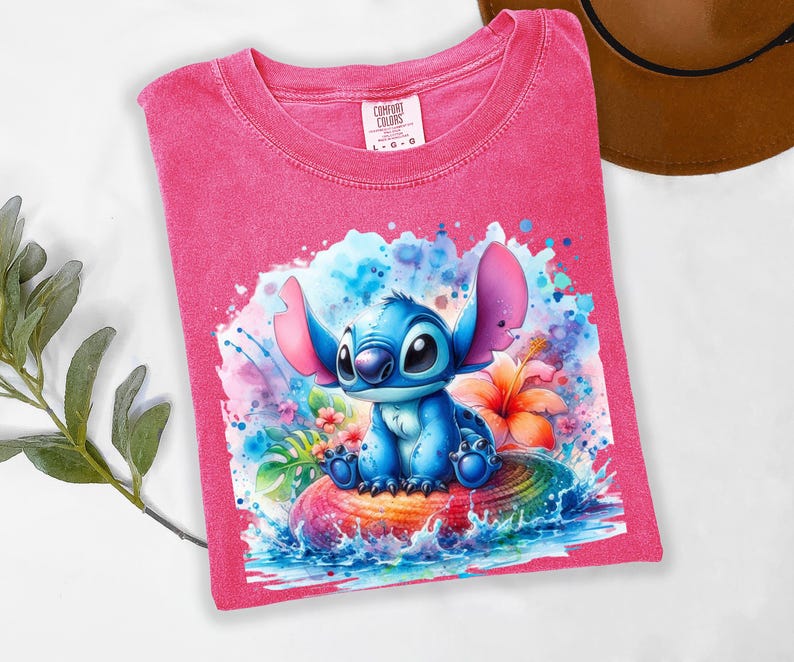 Może przedstawiać: R&oacute;żowa koszulka Comfort Colors z akwarelową ilustracją postaci Stitch. Niebieski Stitch siedzi na kolorowej, tęczowej powierzchni z wodą i kwiatami wok&oacute;ł niego. Koszulka w rozmiarze L-G.