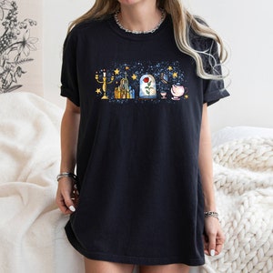 Puede incluir: Una camiseta negra con un estampado gr&aacute;fico de varios elementos ic&oacute;nicos de la pel&iacute;cula de Disney La Bella y la Bestia. El gr&aacute;fico incluye un candelabro, un castillo, una rosa en una c&uacute;pula de vidrio, un libro y una taza de t&eacute;.