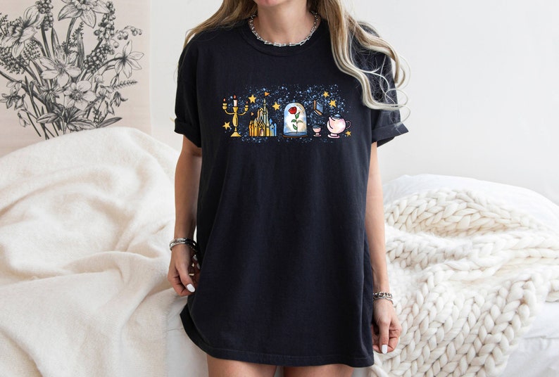 Puede incluir: Una camiseta negra con un estampado gr&aacute;fico de varios elementos ic&oacute;nicos de la pel&iacute;cula de Disney La Bella y la Bestia. El gr&aacute;fico incluye un candelabro, un castillo, una rosa en una c&uacute;pula de vidrio, un libro y una taza de t&eacute;.