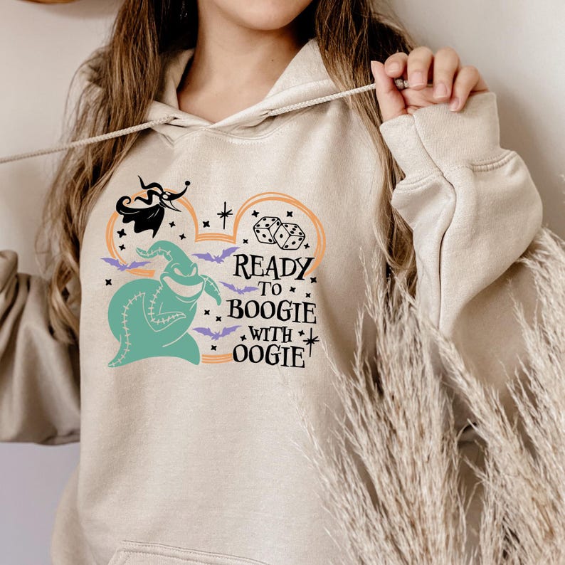 Pu&ograve; includere: Felpa con cappuccio beige con una grafica di Oogie Boogie, pipistrelli, dadi e la scritta "Ready to Boogie with Oogie". Il design include elementi arancioni, verde acqua, neri e viola. La felpa ha un cordino e una tasca frontale.