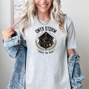 May include: Gray t-shirt with a black and gold graphic design featuring a dragon, moons, and stars. The text "ONYX STORM" and "BRAVE THE DARK" are printed on the shirt.