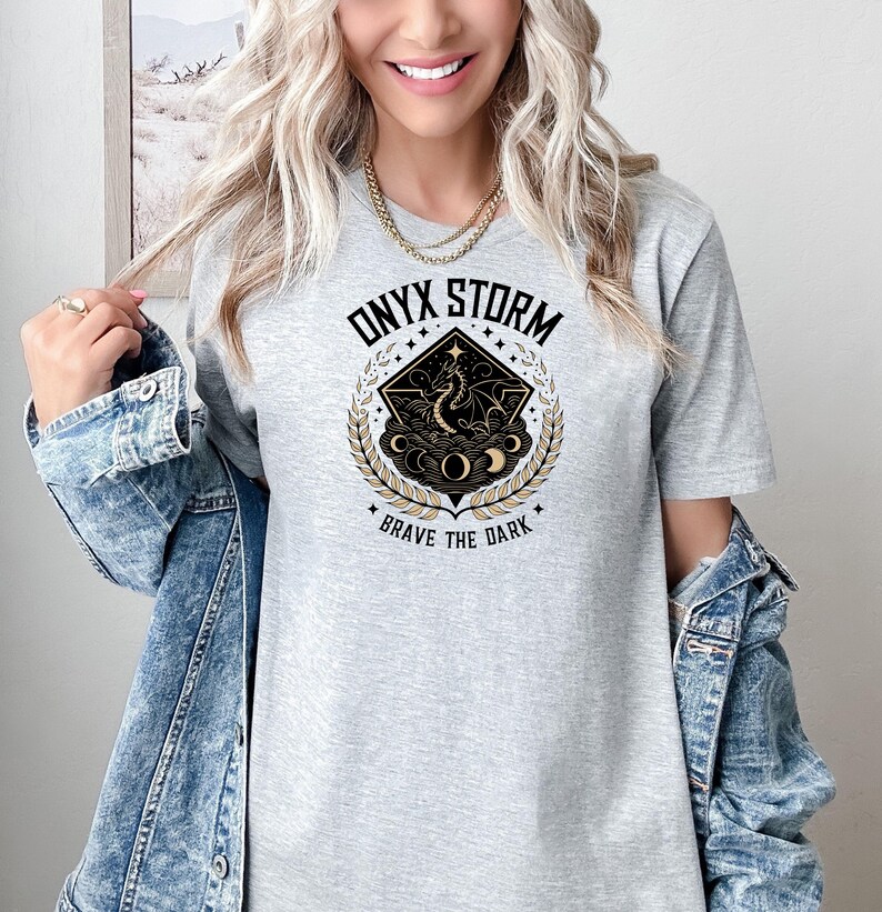 May include: Gray t-shirt with a black and gold graphic design featuring a dragon, moons, and stars. The text "ONYX STORM" and "BRAVE THE DARK" are printed on the shirt.