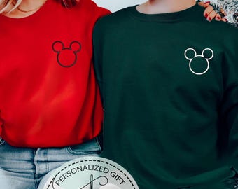 Puede incluir: Una pareja lleva sudaderas rojas y verdes a juego con un diseño de cabeza de Mickey Mouse en blanco y negro en la parte delantera. Las sudaderas son una opción popular para las parejas que buscan una forma divertida y festiva de mostrar su amor por Disney.