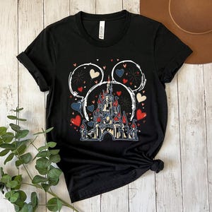 May include: Black t-shirt featuring a castle design with red, white, and blue hearts and fireworks. The design is framed by a white outline of Mickey Mouse ears. A casual, everyday tee.