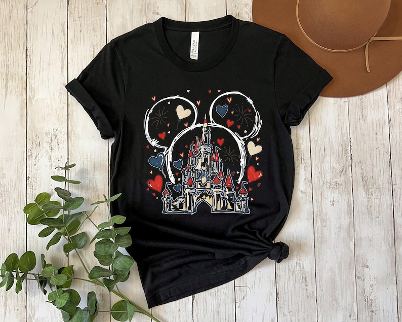 May include: Black t-shirt featuring a castle design with red, white, and blue hearts and fireworks. The design is framed by a white outline of Mickey Mouse ears. A casual, everyday tee.