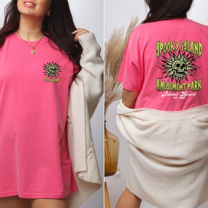 May include: A bright pink t-shirt with a graphic on the front and back. The front graphic is a green sun with a skull and the text "Spooky Island". The back graphic says "Spooky Island Amusement Park Island Resort".