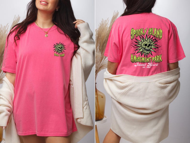 May include: A bright pink t-shirt with a graphic on the front and back. The front graphic is a green sun with a skull and the text "Spooky Island". The back graphic says "Spooky Island Amusement Park Island Resort".