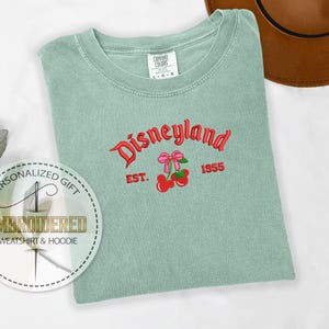 Op de afbeelding: Een lichtgroen T-shirt met de tekst "Disneyland" in rode en zwarte letters. De tekst "Est. 1955" staat onder de Disneyland-tekst. Een rode kers met een roze strik is geborduurd op het shirt.