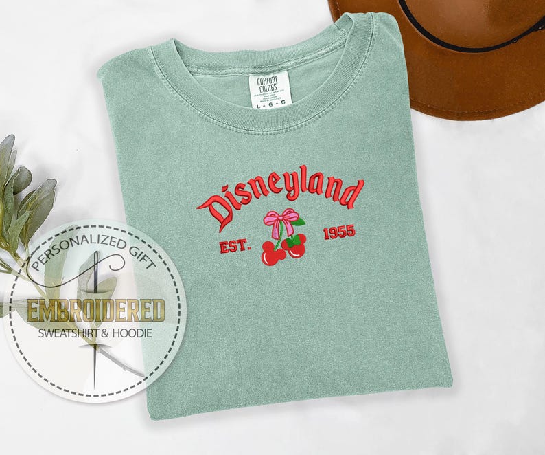 Op de afbeelding: Een lichtgroen T-shirt met de tekst "Disneyland" in rode en zwarte letters. De tekst "Est. 1955" staat onder de Disneyland-tekst. Een rode kers met een roze strik is geborduurd op het shirt.