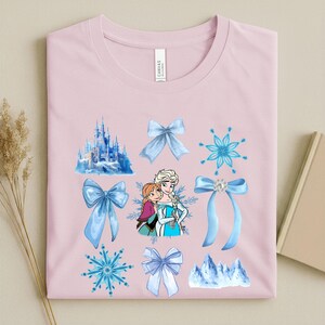 May include: A light pink t-shirt featuring a design with illustrations of a castle, snowflakes, and bows in shades of blue. The shirt also has a graphic of two cartoon characters. The shirt is made of a soft material.