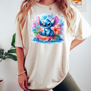 Może przedstawiać: Kremowy t-shirt z kolorową grafiką Stitcha z Lilo & Stitch. Stitch, w kolorze niebieskim, siedzi na nadmuchiwanym kole w kształcie tęczy, otoczony rozpryskami akwareli i tropikalnymi kwiatami. Koszulka ma kr&oacute;tkie rękawy.