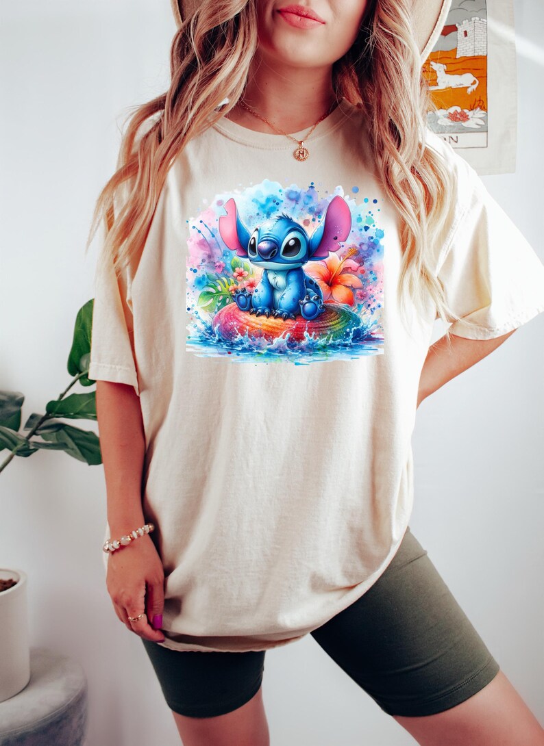 Może przedstawiać: Kremowy t-shirt z kolorową grafiką Stitcha z Lilo & Stitch. Stitch, w kolorze niebieskim, siedzi na nadmuchiwanym kole w kształcie tęczy, otoczony rozpryskami akwareli i tropikalnymi kwiatami. Koszulka ma kr&oacute;tkie rękawy.