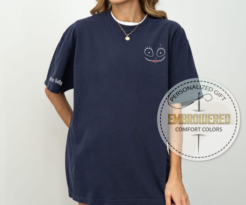 K&ouml;nnte beinhalten: Marineblaues, besticktes Comfort Colors T-Shirt mit einem Cartoon-Gesicht auf der Vorderseite und "His Sally" auf dem &Auml;rmel. Das Design umfasst einen wei&szlig;en Kragen und eine goldene Halskette.