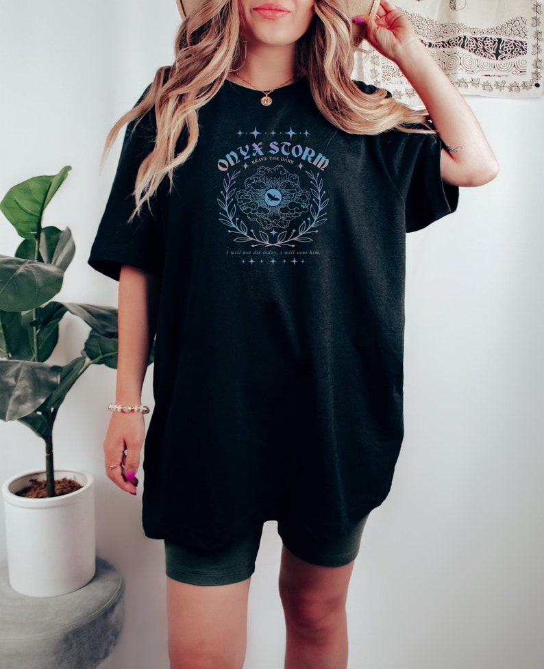 May include: Black oversized t-shirt with a graphic print of a floral design with a blue eye in the center. The text "ONYX STORM" is above the design and "BRAVE THE DARK" is below the design. The text "I will not die today, I will save him." is at the bottom of the graphic.