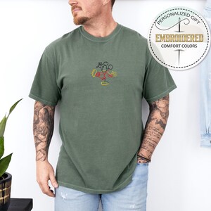 May include: A green t-shirt with a simple embroidered design of Mickey Mouse. The design is in red, yellow, and black. The t-shirt is a comfortable, casual style.