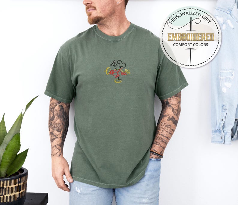 May include: A green t-shirt with a simple embroidered design of Mickey Mouse. The design is in red, yellow, and black. The t-shirt is a comfortable, casual style.