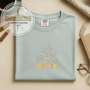 May include: A light blue t-shirt with an embroidered castle design and the words "Disney World" in gold. The shirt has a crew neck and a tag that says "Comfort Colors". The image also includes a personalized gift tag.