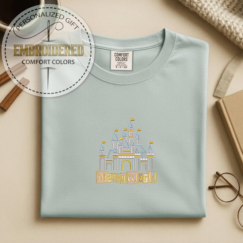 May include: A light blue t-shirt with an embroidered castle design and the words "Disney World" in gold. The shirt has a crew neck and a tag that says "Comfort Colors". The image also includes a personalized gift tag.