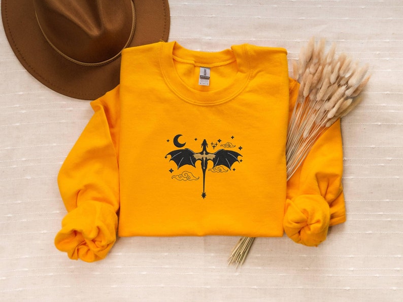 May include: A yellow sweatshirt featuring a black dragon graphic with a crescent moon, stars, and clouds. The dragon is flying with its wings outstretched.